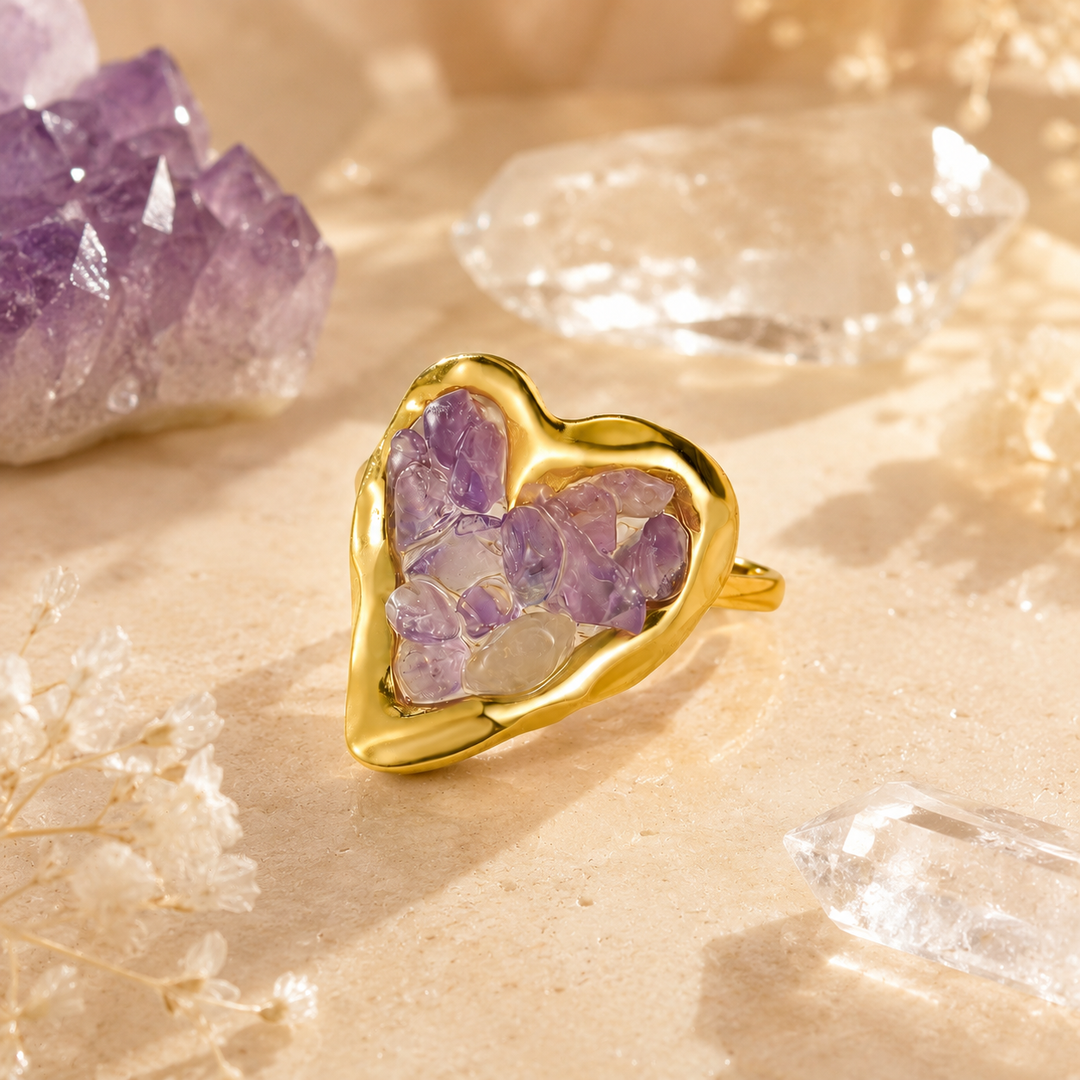 Gold-tone heart-shaped ring with amethyst crystal chips displayed on a warm stone surface with clear quartz and purple crystal background elements.