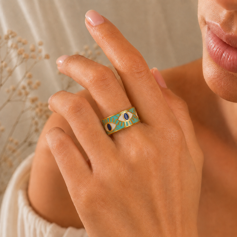 Orphaya turquoise evil eye ring worn on hand, featuring a wide gold-tone adjustable band with turquoise enamel, navy eye details, and a warm soft-lit lifestyle jewelry background.