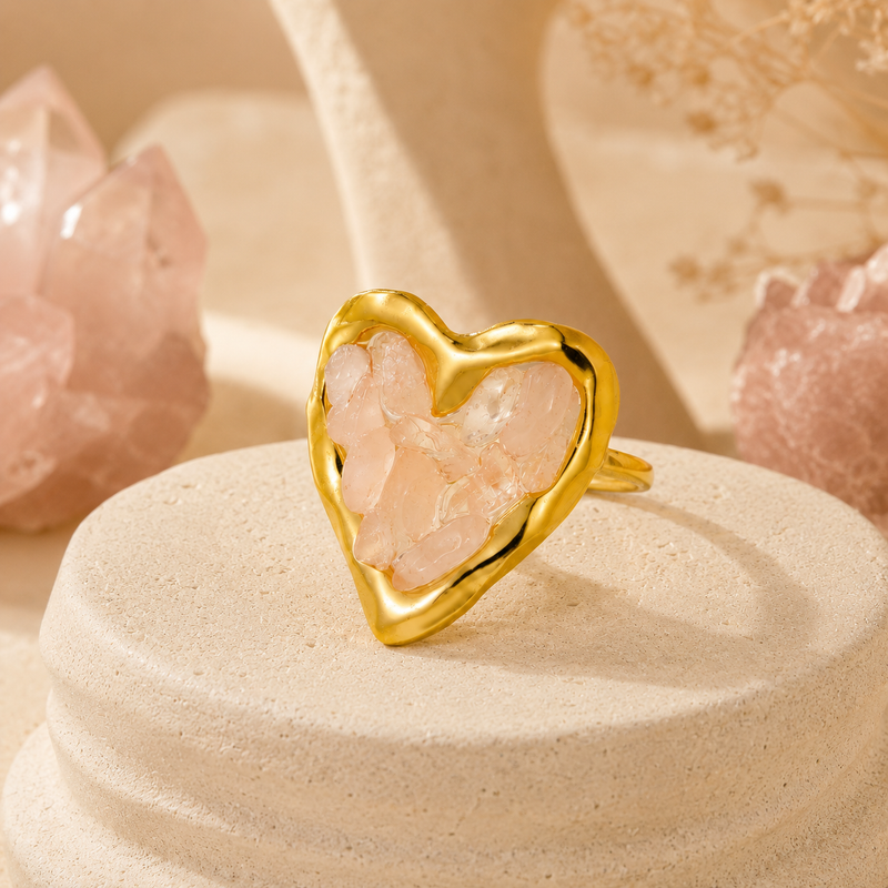 Orphaya gold heart ring with soft pink rose quartz crystal chips displayed on a warm neutral stone background.